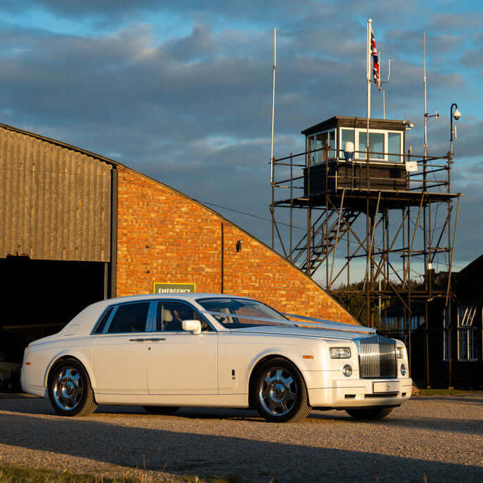 Elegant white Rolls-Royce Phantom parked outside an aviation hangar at sunset, symbolising timeless luxury and sophistication. Experience VIP chauffeur hire with SJ Chauffeurs, specialising in weddings, executive travel, and luxury event transport across the UK. Book your Rolls-Royce Phantom chauffeur today!