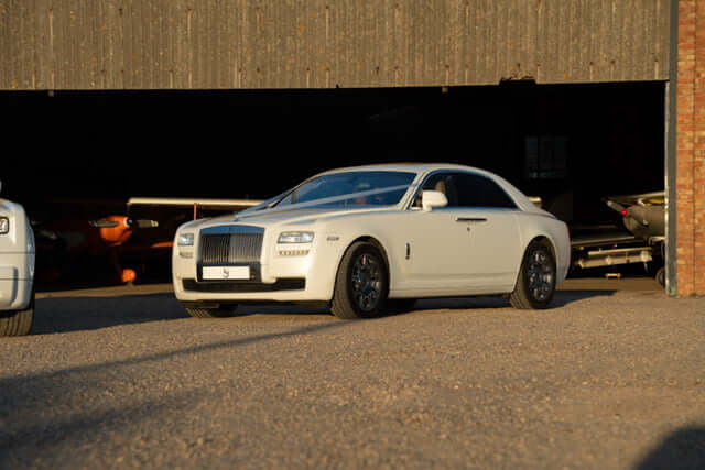 Stunning white Rolls-Royce Ghost parked outside an aircraft hangar, symbolizing luxury and exclusivity. Elevate your travel experience with SJ Chauffeurs, providing VIP, executive, and wedding chauffeur services across the UK. Arrive in elegance—book your Rolls-Royce chauffeur today!